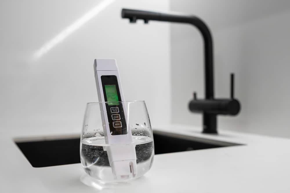 Digitaler Wassertester in einem Glas Wasser vor einem modernen schwarzen Wasserhahn in einer minimalistischen Küche.