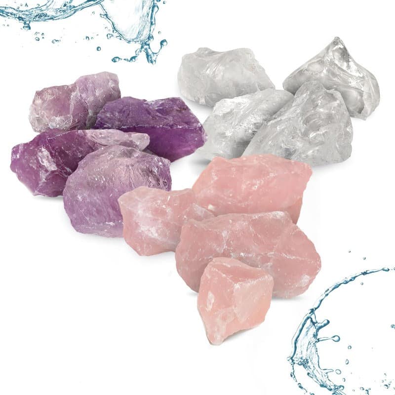 Rohsteine in den Farben Lila, Rosa und Transparent, umgeben von Wasser. Das Bild zeigt Amethyst, Rosenquarz und Bergkristall in natürlicher, unbearbeiteter Form mit spritzendem Wasser im Hintergrund.