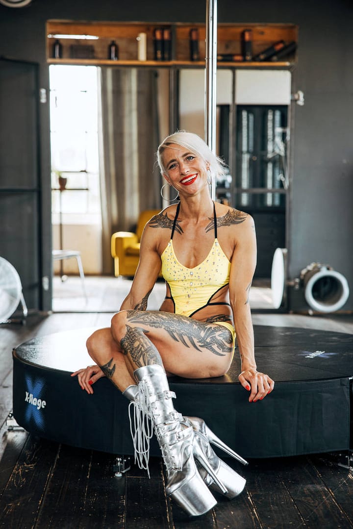Die Poledance-Tänzerin Éowyn Eichhorn in einem gelben Kostüm und silbernen Plateau-Stiefeln sitzt lächelnd auf einer mobilen Poledance-Bühne in einem Tanzstudio. Ihr Körper ist mit auffälligen Tätowierungen verziert.